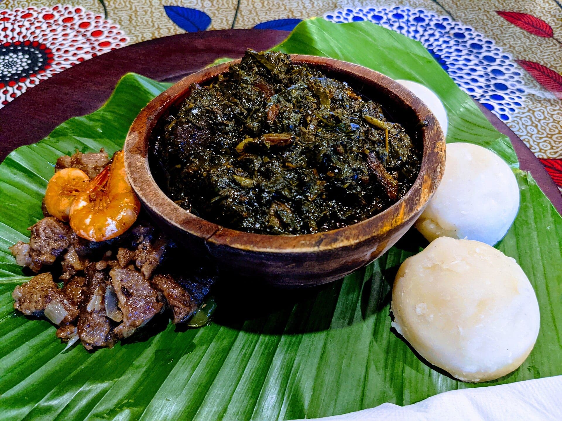 Un bol d’eru entouré de boules de water fufu et de viande, sur une feuille