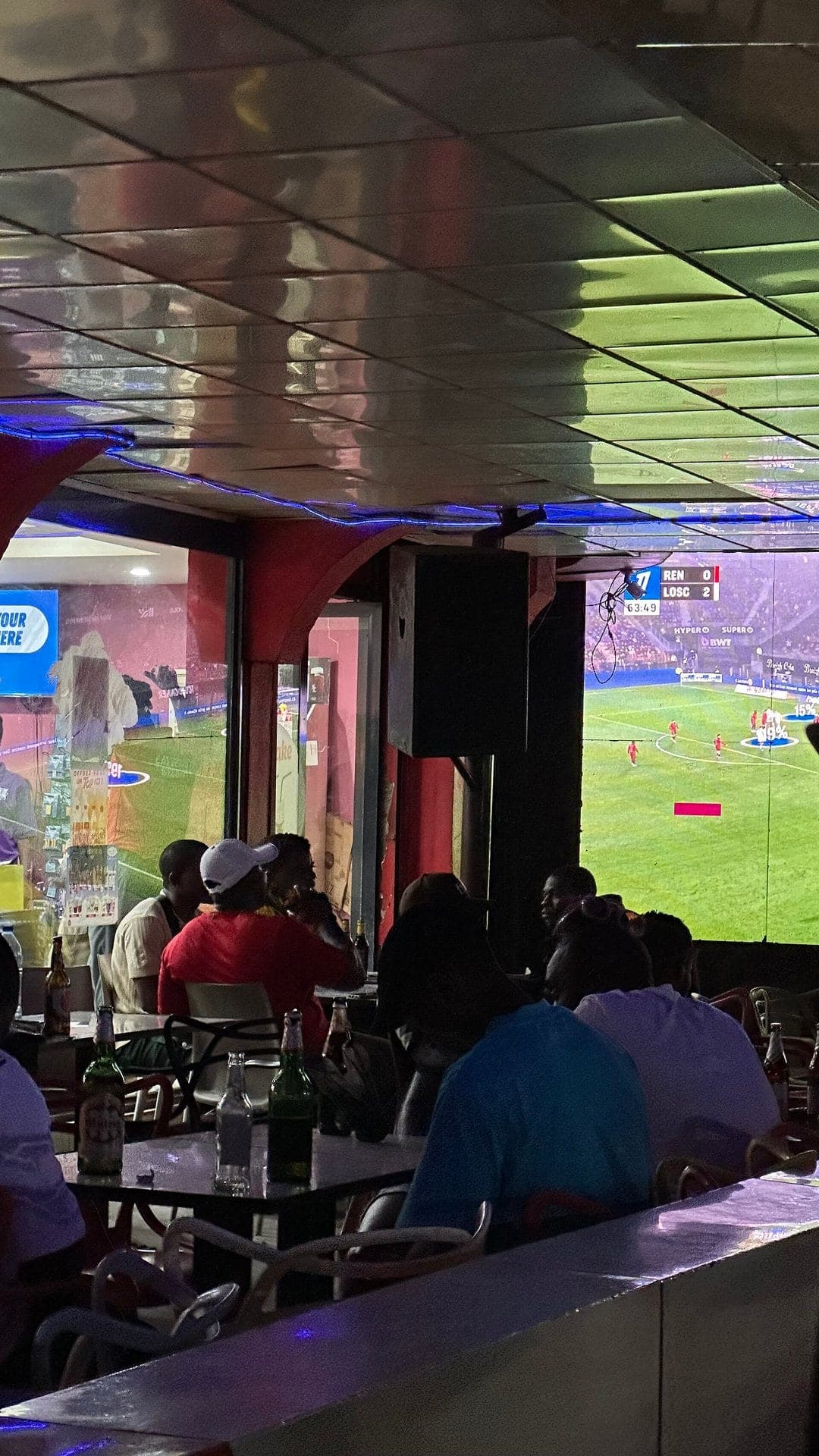 Des clients attablés, de dos, regardent un match sur grand écran dans un bar