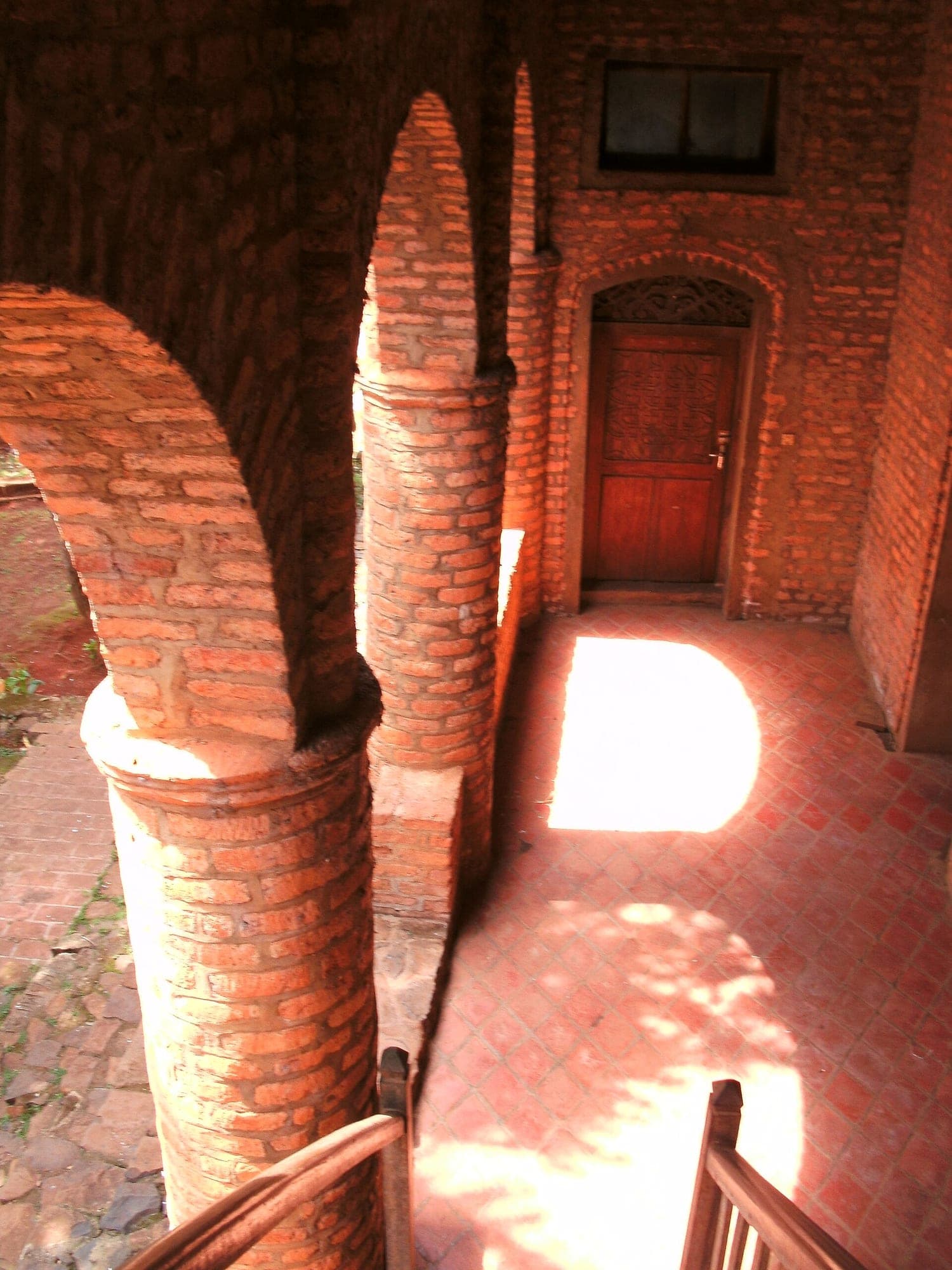Des arcades et des colonnes de brique rouge dans le palais de Foumban, traversées par la lumière